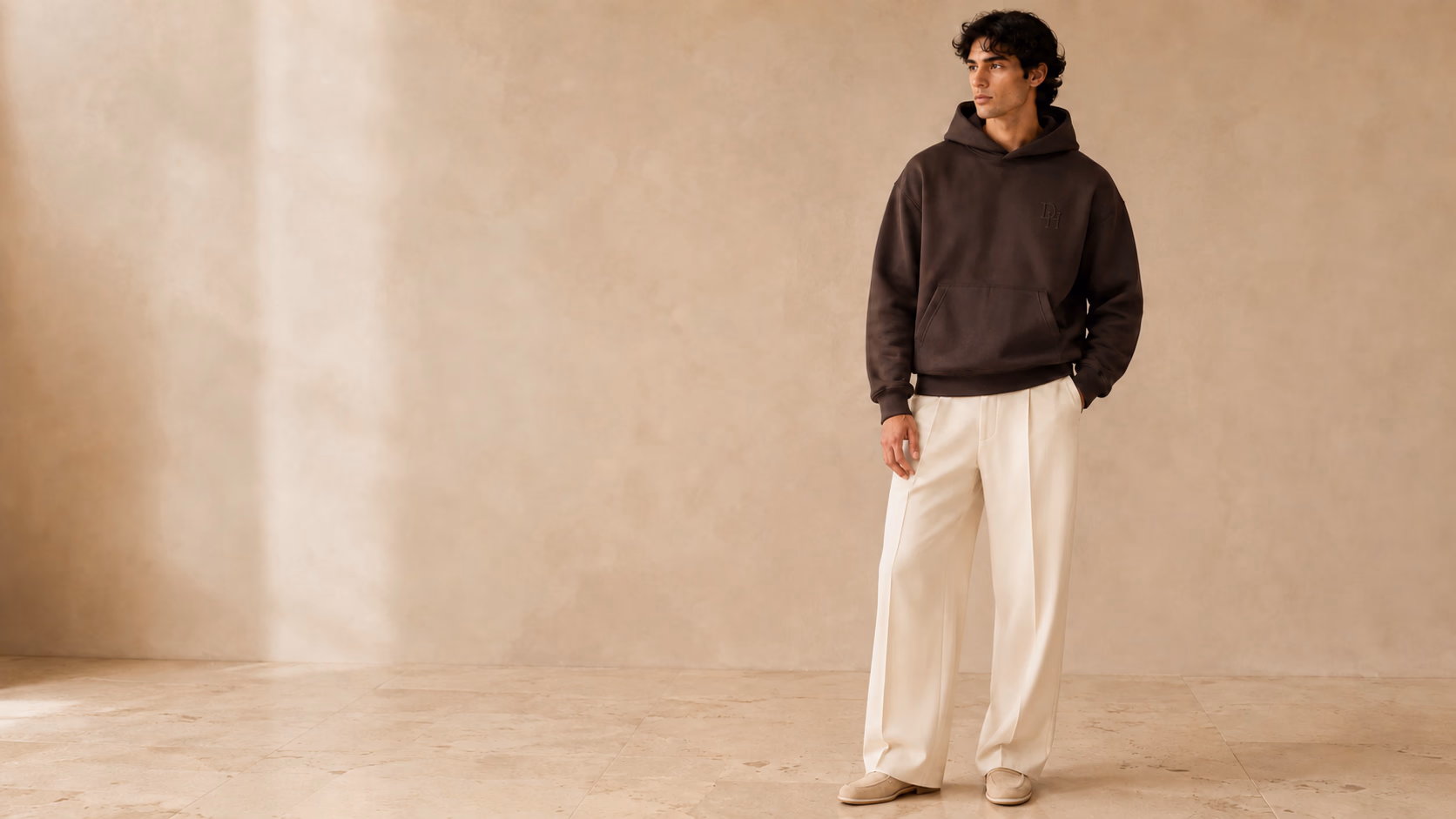 Model wearing the stringless espresso Dynast Heir heavyweight hoodie with tonal embroidered DH monogram and cream tailored trousers